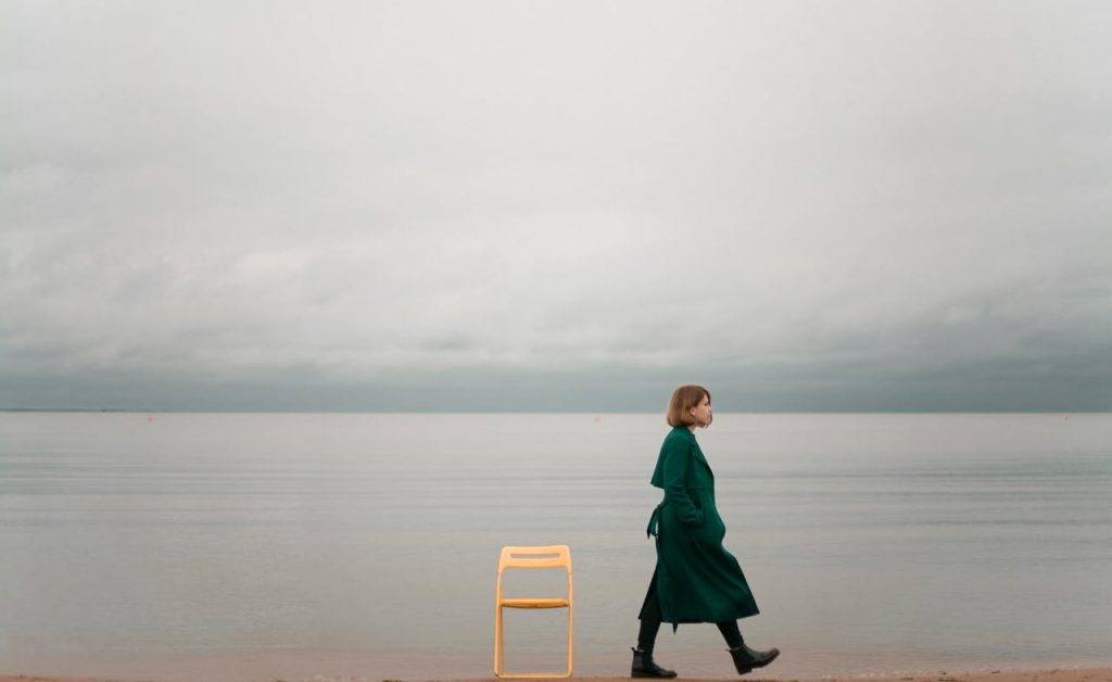 A woman in a green coat walking along a calm seaside beside a yellow chair under a cloudy sky.