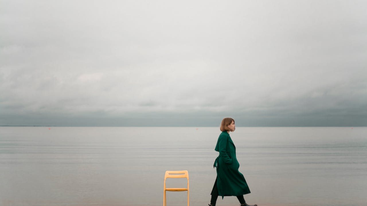 A woman in a green coat walking along a calm seaside beside a yellow chair under a cloudy sky.