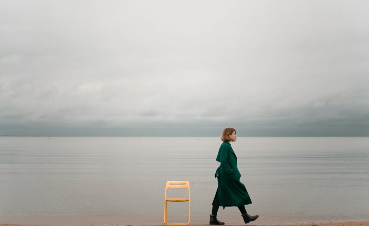 A woman in a green coat walking along a calm seaside beside a yellow chair under a cloudy sky.