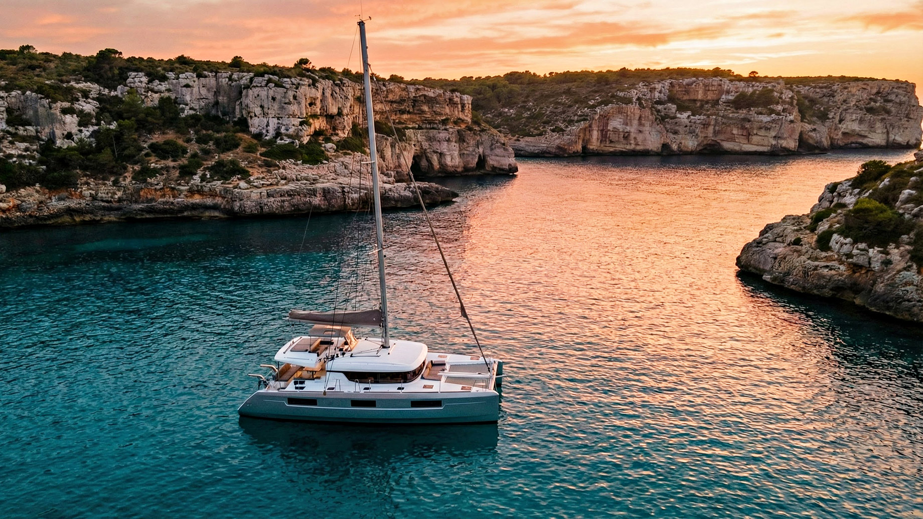 Aerial view of a white catamaran sailboat anchored in a calm, shallow bay in Mallorca, Spain. The vessel is completely empty with no passengers on deck or in the surrounding crystal-clear turquoise water, set against a backdrop of rocky coastline and green vegetation.
