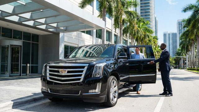 A professional chauffeur in a suit standing beside a sleek black luxury sedan on a city street, representing reliable executive limousine services for long-distance and metropolitan travel.