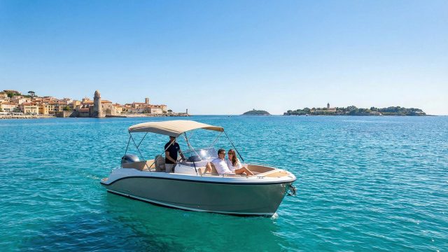 A couple relaxes on the bow of a small motorboat with a skipper at the helm, cruising on turquoise waters. In the background is the coastal town of Cannes with a prominent stone tower and an island under a clear blue sky.