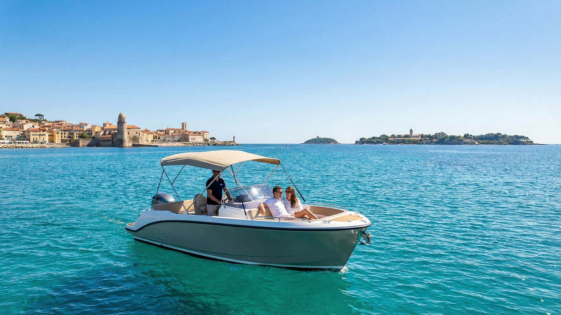 A couple relaxes on the bow of a small motorboat with a skipper at the helm, cruising on turquoise waters. In the background is the coastal town of Cannes with a prominent stone tower and an island under a clear blue sky.