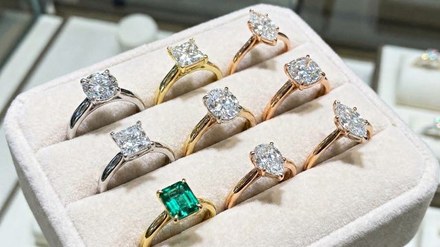 A close-up photograph of a jewelry display box lined with light beige velvet, showcasing nine engagement rings. The rings feature various diamond cuts, including round brilliant, princess, oval, marquise, and pear shapes, set in white gold, yellow gold, and rose gold bands. One ring in the foreground features a rectangular green emerald gemstone. The background is slightly blurred but suggests a jewelry store setting.