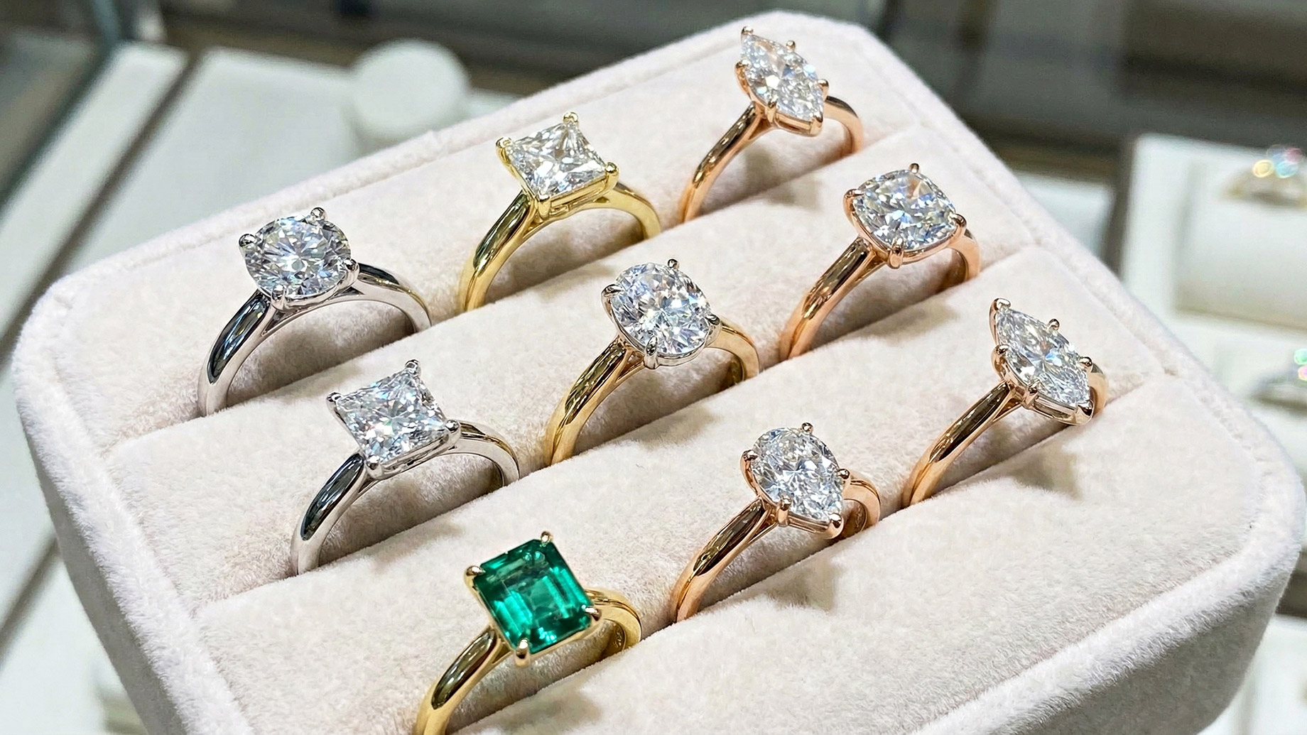 A close-up photograph of a jewelry display box lined with light beige velvet, showcasing nine engagement rings. The rings feature various diamond cuts, including round brilliant, princess, oval, marquise, and pear shapes, set in white gold, yellow gold, and rose gold bands. One ring in the foreground features a rectangular green emerald gemstone. The background is slightly blurred but suggests a jewelry store setting.