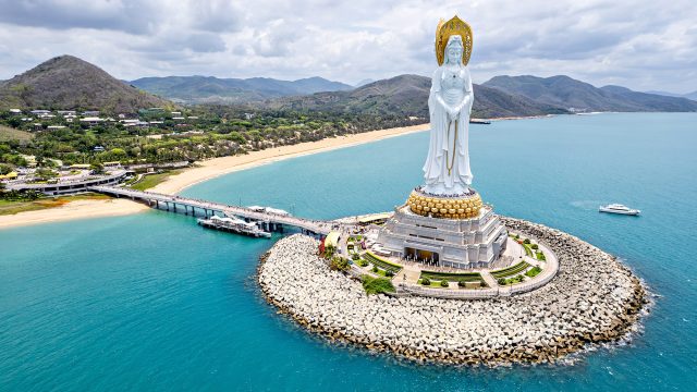Aerial view of the Guanyin of Nanshan statue rising above turquoise water, sandy beaches, and mountain scenery in Sanya.