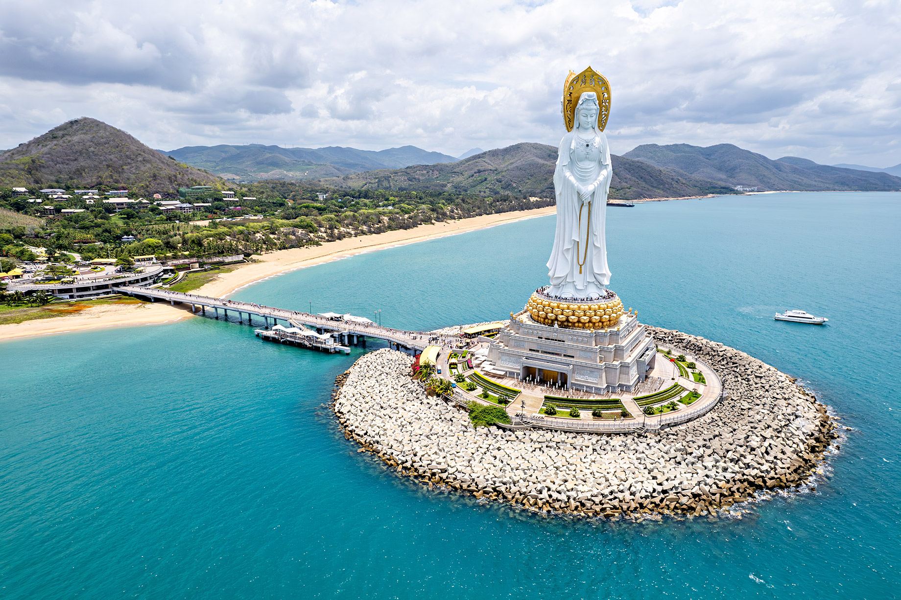 Aerial view of the Guanyin of Nanshan statue rising above turquoise water, sandy beaches, and mountain scenery in Sanya.