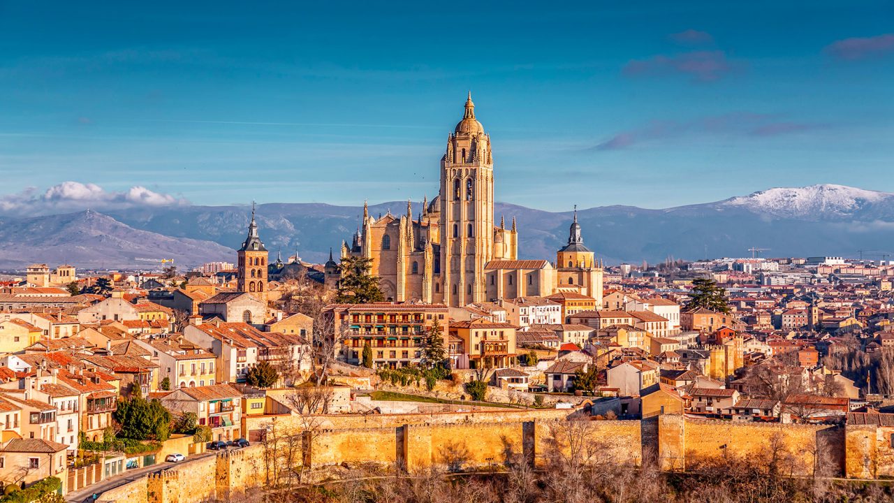 Segovia Cathedral rising above the terracotta rooftops and medieval walls of Segovia, with snow-covered mountains in the distance.