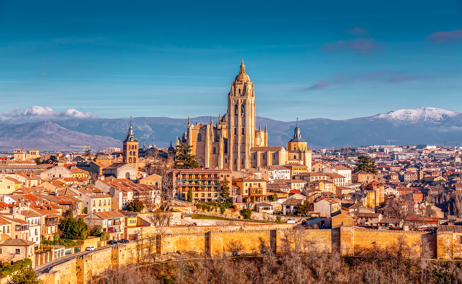 Segovia Cathedral rising above the terracotta rooftops and medieval walls of Segovia, with snow-covered mountains in the distance.