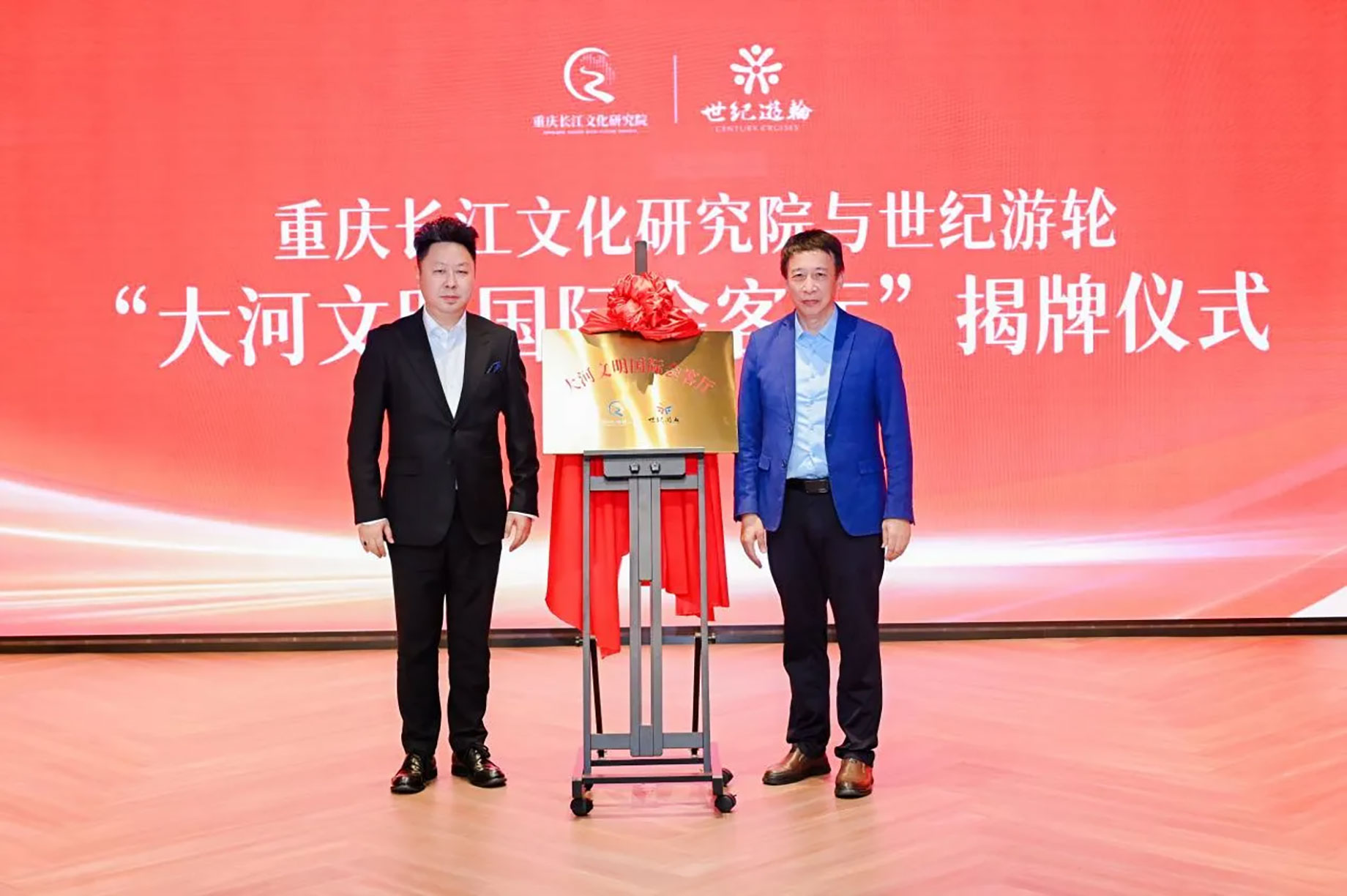 Two men stand beside a gold plaque topped with a red bow at a Century Cruises unveiling ceremony.