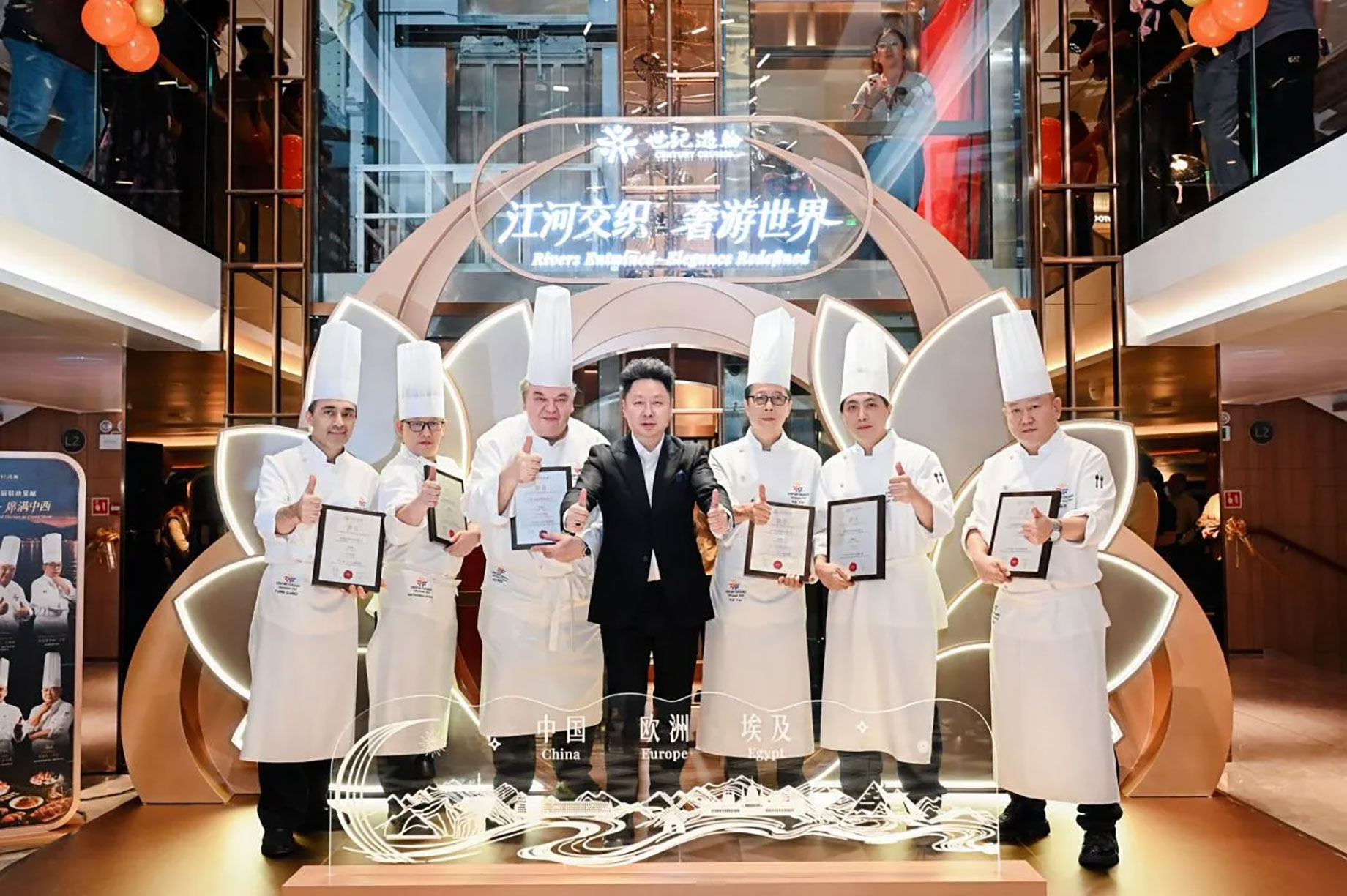 Six chefs in white uniforms hold framed certificates beside a suited man in an atrium decorated with an illuminated floral display.