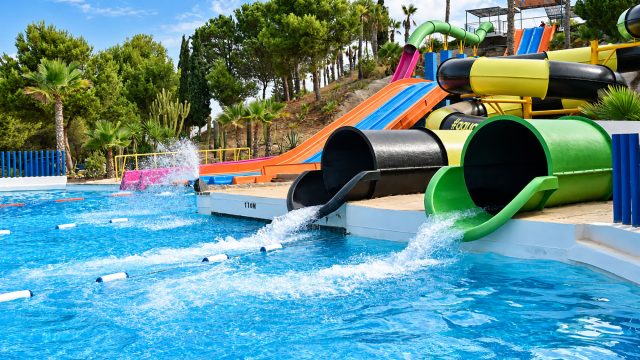 Colorful open and enclosed water slides flowing into a bright blue resort pool surrounded by palm trees and tropical landscaping.