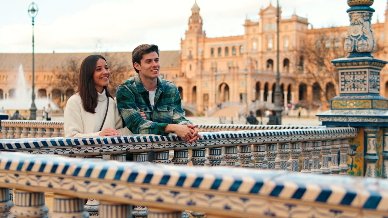 Smiling couple leaning on a blue-and-white tiled bridge railing at Plaza de España in Seville, with brick arcades behind them.