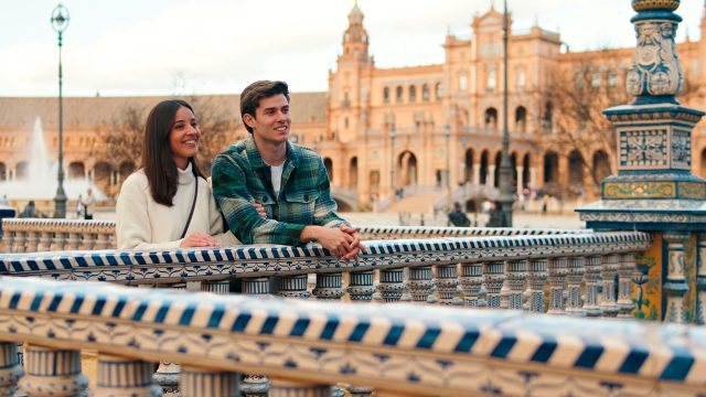 Smiling couple leaning on a blue-and-white tiled bridge railing at Plaza de España in Seville, with brick arcades behind them.