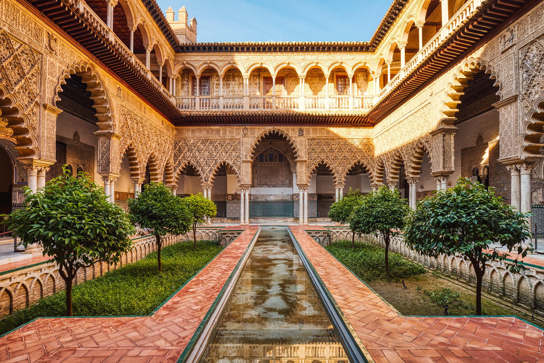 A long reflecting pool between garden beds and ornate arched galleries in a courtyard at the Real Alcázar in Seville, Spain.