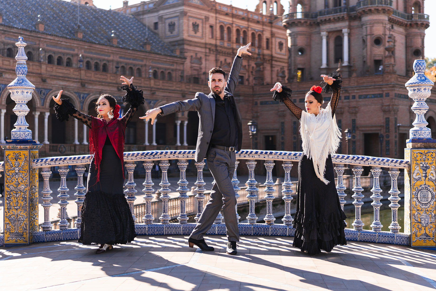 Three flamenco dancers with raised arms on a blue and white tiled bridge at Plaza de España in Seville, Spain.