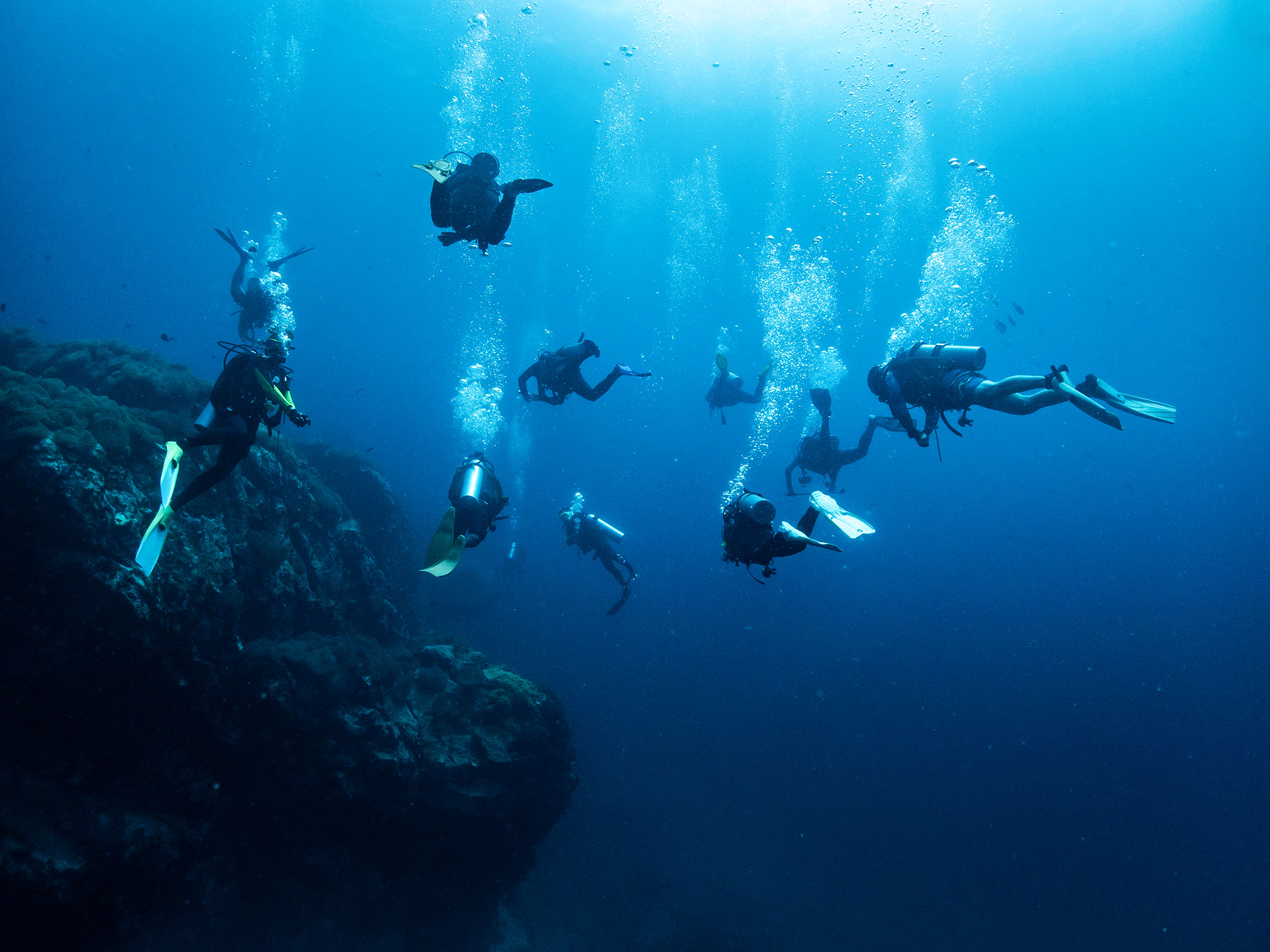 Scuba divers swim beside a rocky reef at Koh Tao, Thailand, with streams of bubbles rising through the blue water.