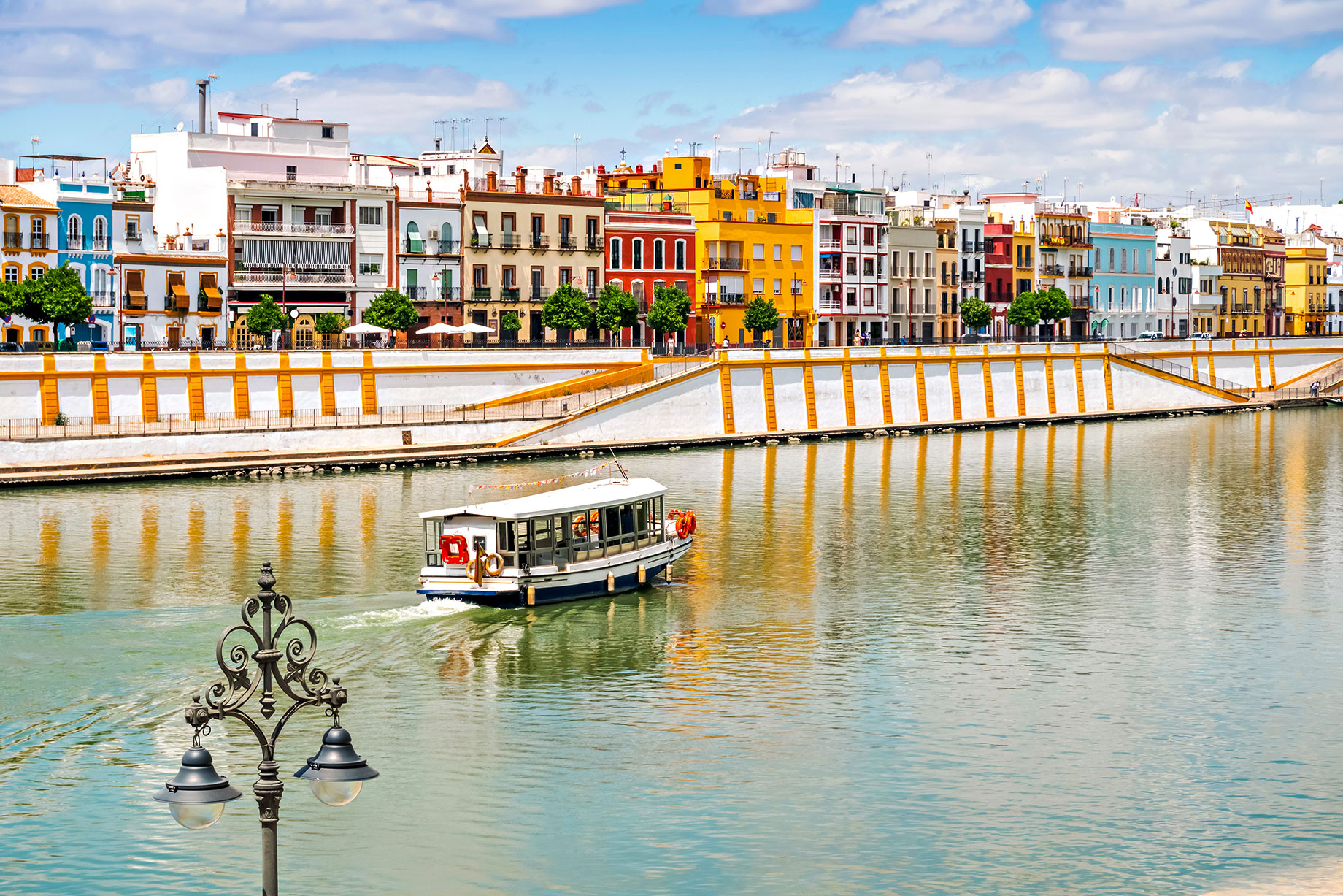 A sightseeing boat passes colourful waterfront buildings along the Guadalquivir River in Seville, Spain.