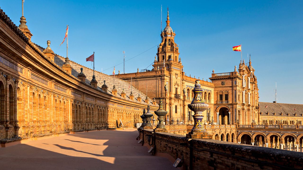 Sunlit brick arcades and ornate ceramic finials along an upper terrace at Plaza de España in Seville, Spain.