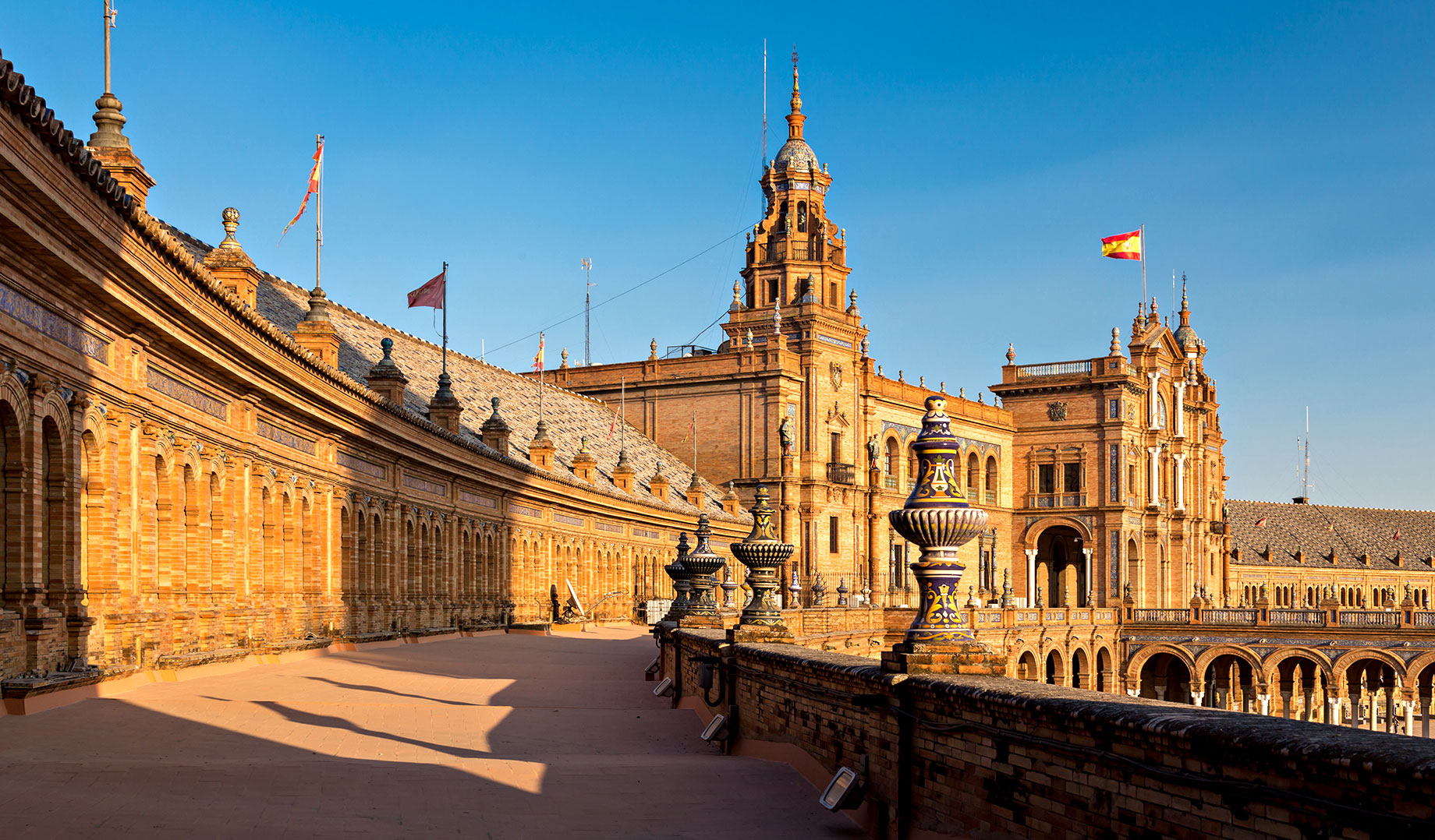 Sunlit brick arcades and ornate ceramic finials along an upper terrace at Plaza de España in Seville, Spain.