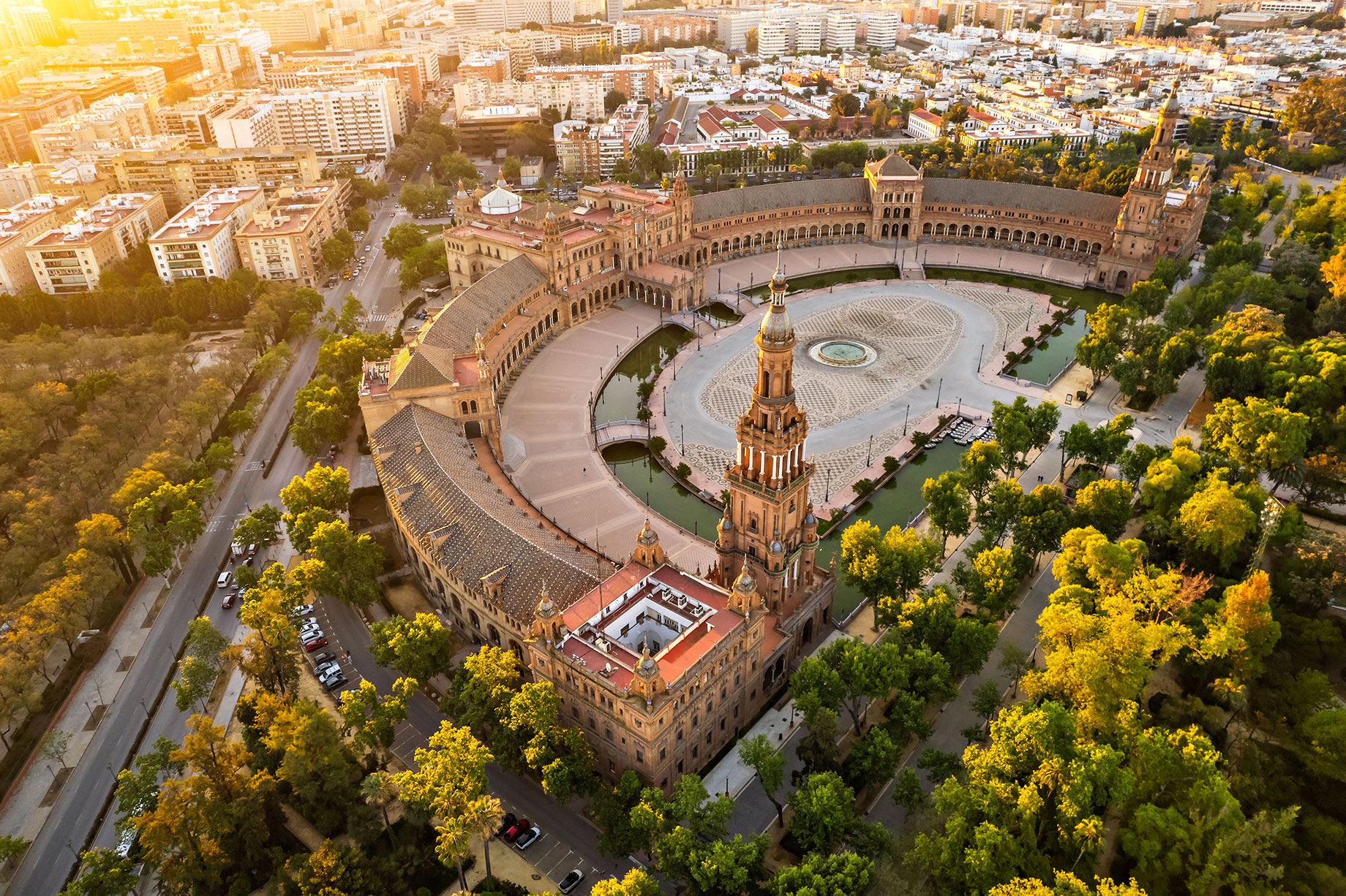 Aerial view of Plaza de España’s towers, curved canal and arcades beside María Luisa Park in Seville at sunrise.