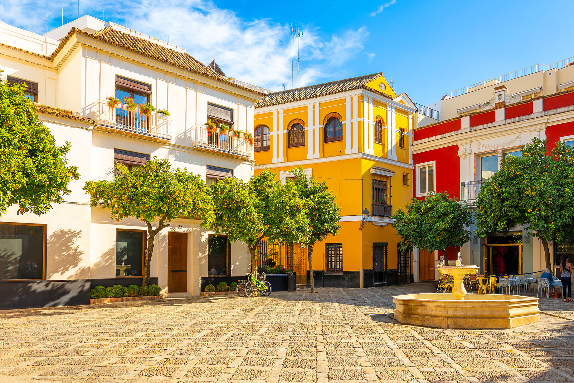 A stone fountain, orange trees and café tables surrounded by colourful façades in Plaza de la Alianza, Santa Cruz, Seville.