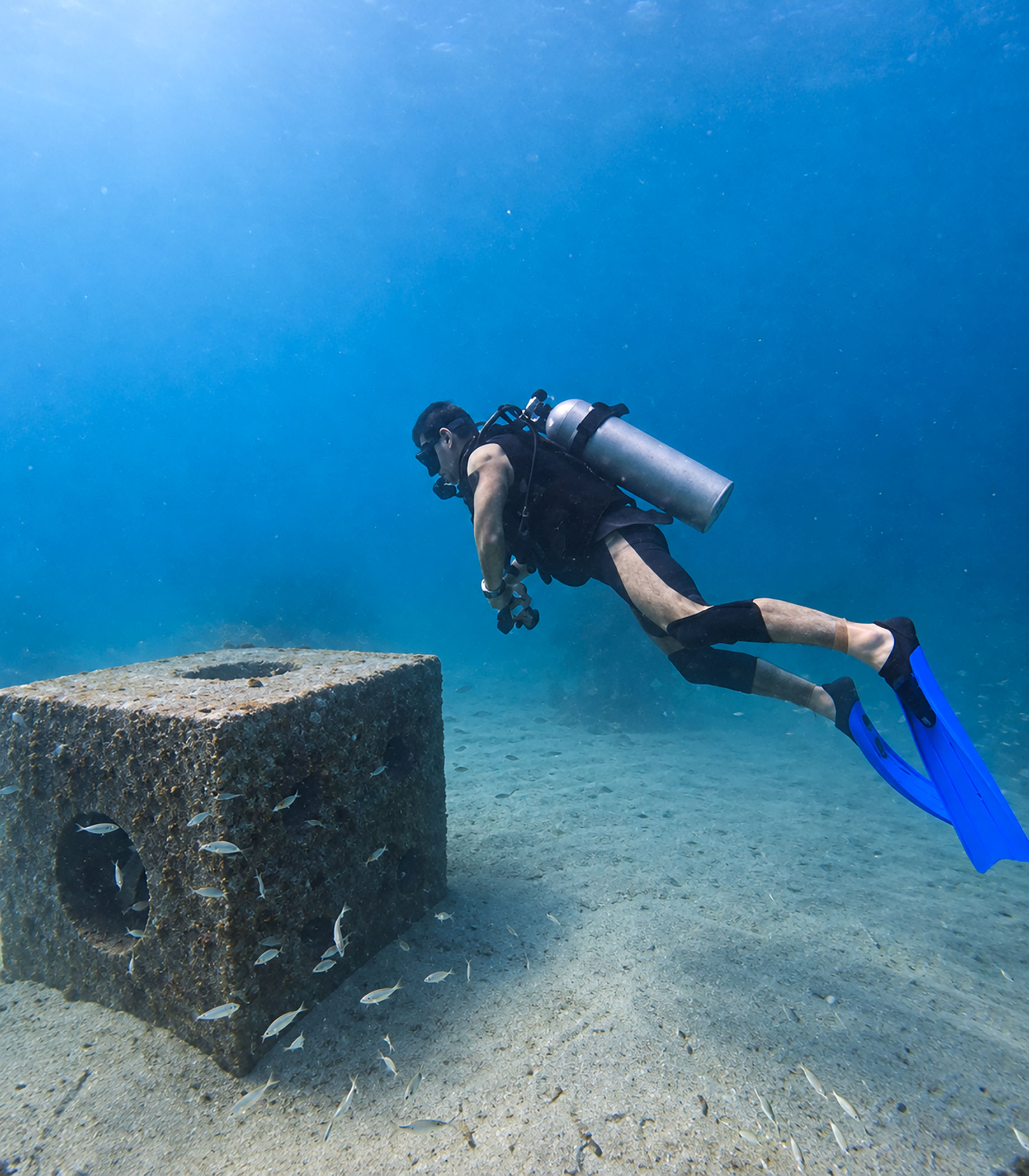 A scuba diver with blue fins swims beside a submerged structure and small fish at the Old Cement Barge dive site in Abu Dhabi.