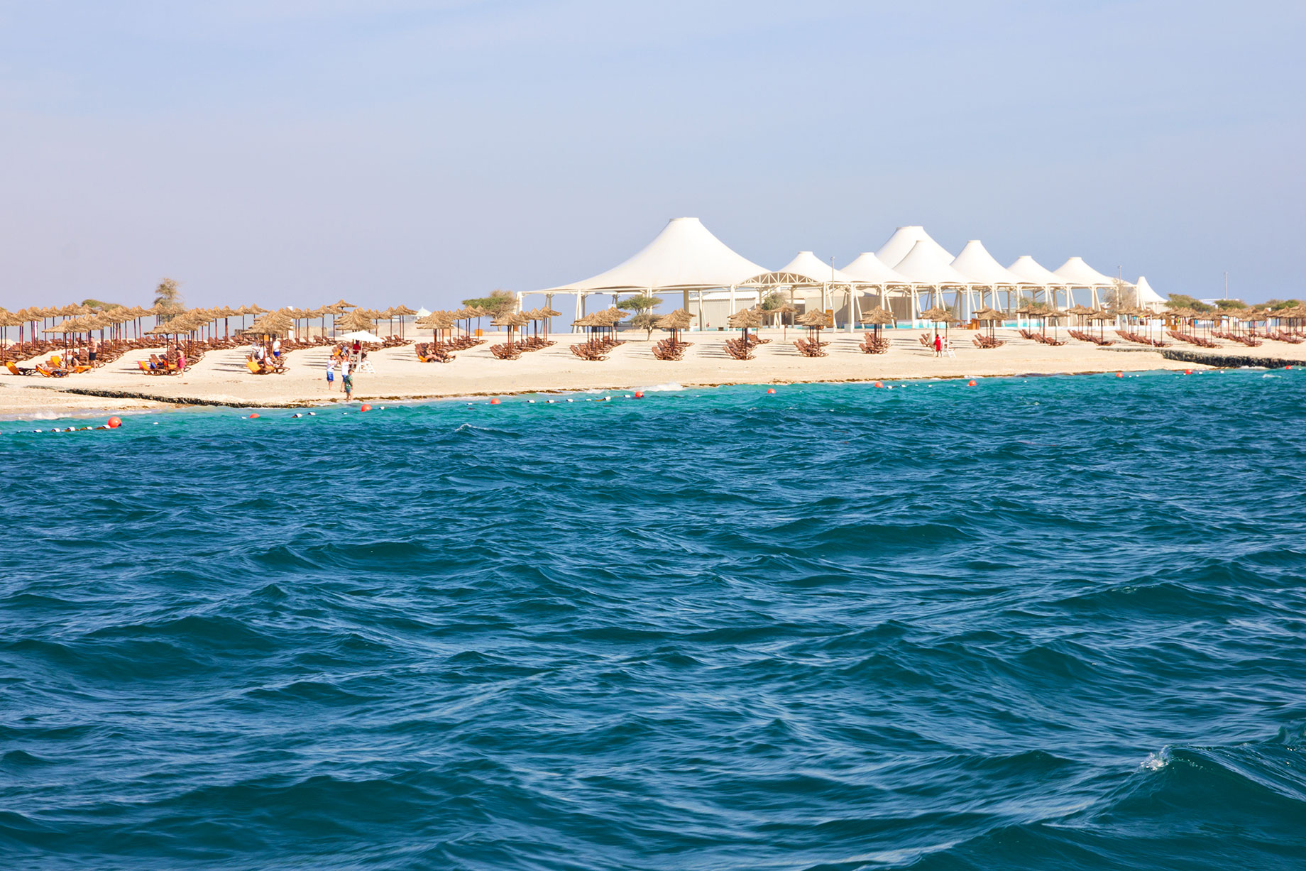 Sandy beach on Sir Bani Yas Island with thatched parasols, sun loungers and white canopy roofs, viewed across turquoise water.