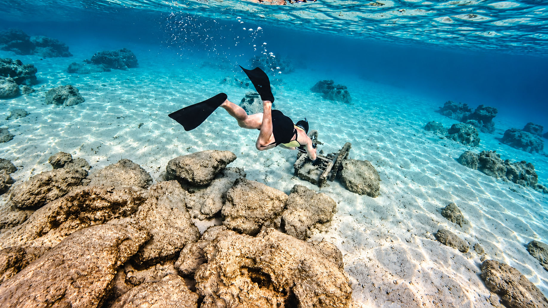 A snorkeller with black fins explores a sandy seabed among scattered rocks in the shallow turquoise waters of Jubail Island.