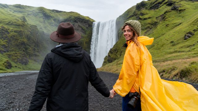 Two travellers hold hands in front of an Icelandic waterfall, one wearing a yellow raincoat and the other a black jacket and brown hat.