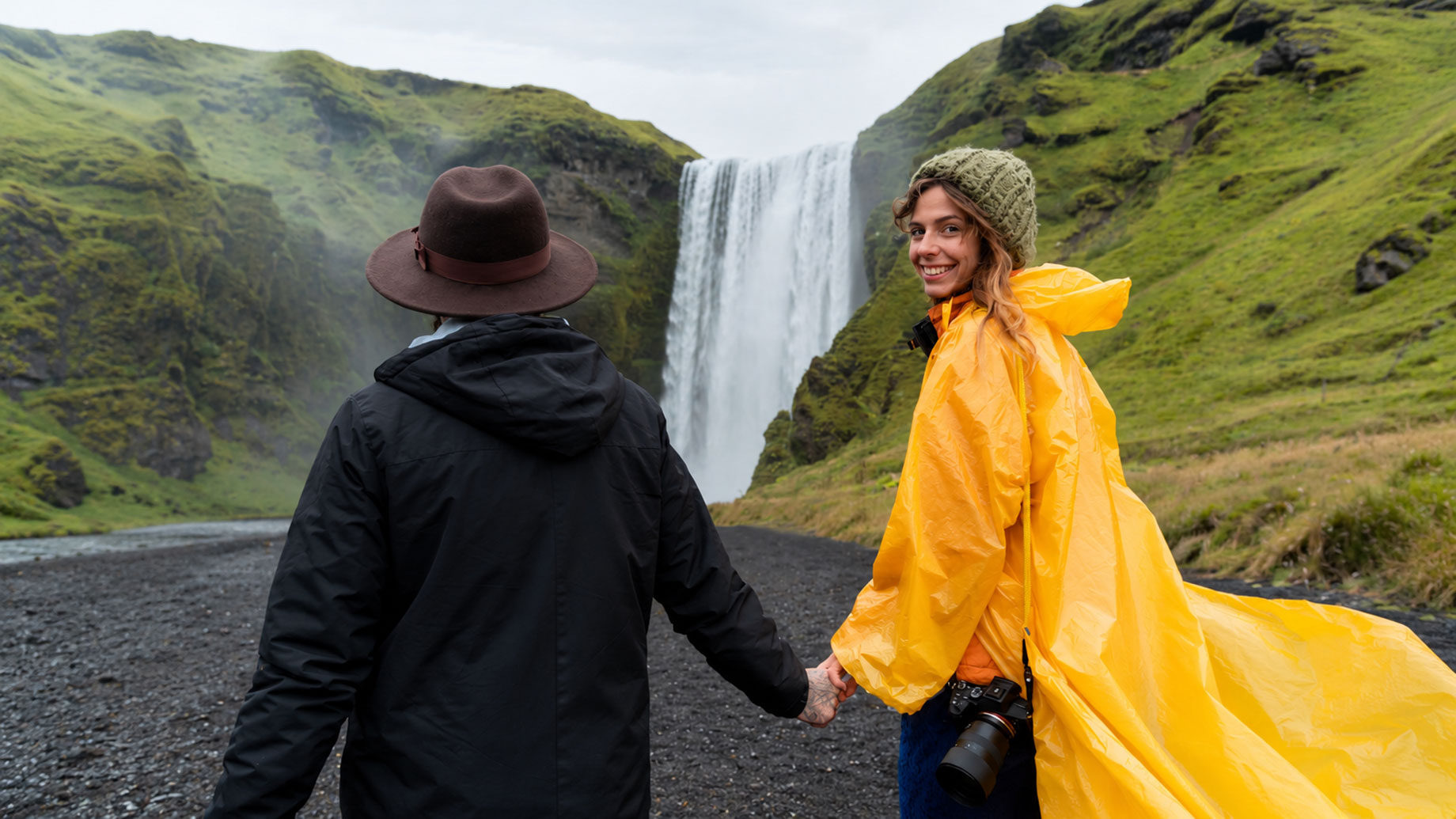 Two travellers hold hands in front of an Icelandic waterfall, one wearing a yellow raincoat and the other a black jacket and brown hat.